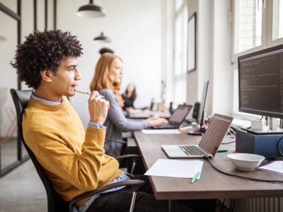 Side view shot of young man sitting at his desk looking at desktop screen and thinking with colleagues working in background. Male computer programmer thinking over new software development in office.