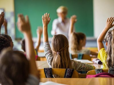 Rear view of large group of students raising their arms to answer the question on a class at elementary school.