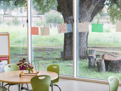A tidy preschool or kindergarten classroom is prepared for students. A large window wall looks out on a large tree and tree stump.