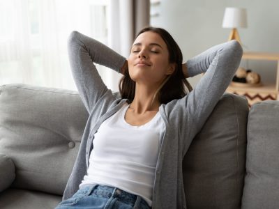 Relaxed serene pretty young woman feel fatigue lounge on comfortable sofa hands behind head rest at home, happy calm lady dream enjoy wellbeing breathing fresh air in cozy home modern living room