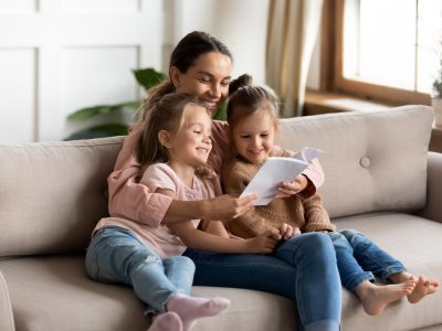 Happy family young mother babysitter hold read book relax embrace cute little children daughters, smiling parent mum tell small kids funny fairy tale story sit on sofa having fun together at home