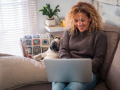 Cheerful and nice couple with people and animal - beautiful culry adult woman with funny dog pug at home using personal computer laptop sitting on the sofa together looking the screen