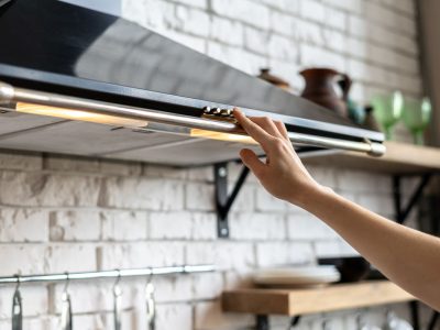 Kitchen with modern interior, furniture and equipment Cropped view of woman hand select mode on cooking hood, standing near kitchen appliance in contemporary interior with brick wall and decor on shelves at blurred background