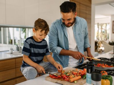 Little boy cutting vegetables while his father cooking food in kitchen. Father and son preparing food at home kitchen.